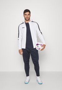 Witte zip-up jack met zwarte zijstrepen, marineblauw T-shirt, marineblauwe trainingsbroek en witte schoenen, terwijl hij een voetbal met een kleurrijk ontwerp vasthoudt.