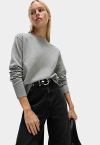 Grauer Pullover mit rundem Ausschnitt und gerippten Bündchen, kombiniert mit hoch taillierter schwarzer Jeans und einem schwarzen Gürtel mit glänzender Schnalle.