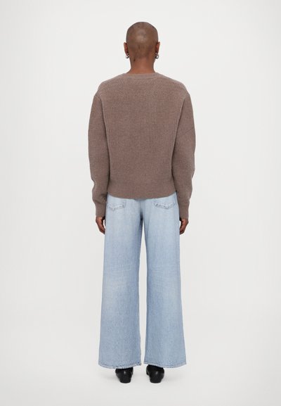 Brauner Strickpullover mit gerippter Textur und weiten Ärmeln, kombiniert mit hellblauen, weit geschnittenen Jeans, die an den Knöcheln enden.