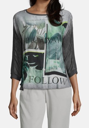 Camisa gráfica gris y verde de mangas 3/4, con un diseño de impresión mixto de hojas y texto. Textura suave y corte casual.