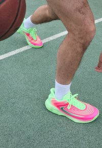 Scarpe da atletica neon verdi e rosa con motivi a onda, materiale testurizzato e suola ammortizzata. Indossate con calze bianche fino al ginocchio su un campo.