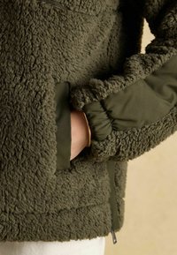 Kurtka z polaru w oliwkowozielonym kolorze o teksturowanej powierzchni, z kontrastującym gładkim materiałem na mankietach oraz zapinaną na zamek kieszenią po boku.