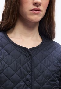 Νεαρή γυναίκα με ανοιχτόχρωμο δέρμα και καστανά μαλλιά, που φοράει μαύρο καπιτονέ μπουφάν με κουμπιά, εμφανιζόμενη από τον λαιμό ως το πάνω μέρος του στήθους.