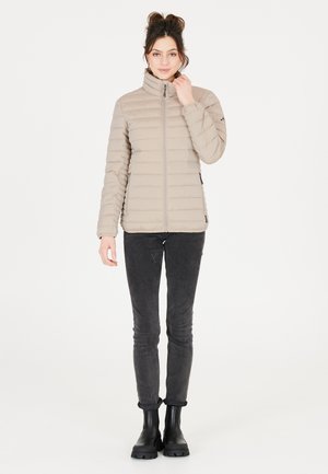 Beige pufferjack met horizontale gestikte patroon, hoge kraag, ritssluiting aan de voorkant en zijzakken. Draagt over een donkere jeans en zwarte laarzen.