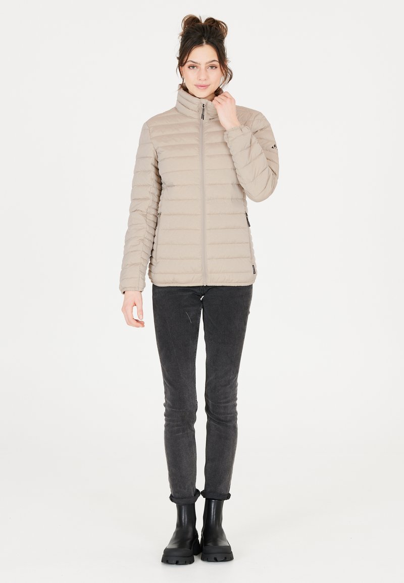 Beige pufferjack met horizontale gestikte patroon, hoge kraag, ritssluiting aan de voorkant en zijzakken. Draagt over een donkere jeans en zwarte laarzen.