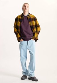 Plaid flannel shirt in yellow and black, purple sweatshirt, light blue jeans, and gray sneakers. Model stands upright against a light background.