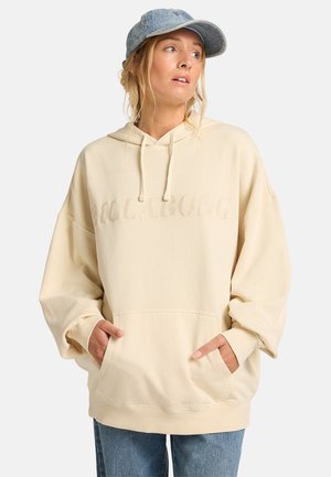 Cream-colored oversized hoodie with a textured logo, drawstring hood, and kangaroo pocket. Paired with light blue jeans and a denim cap.