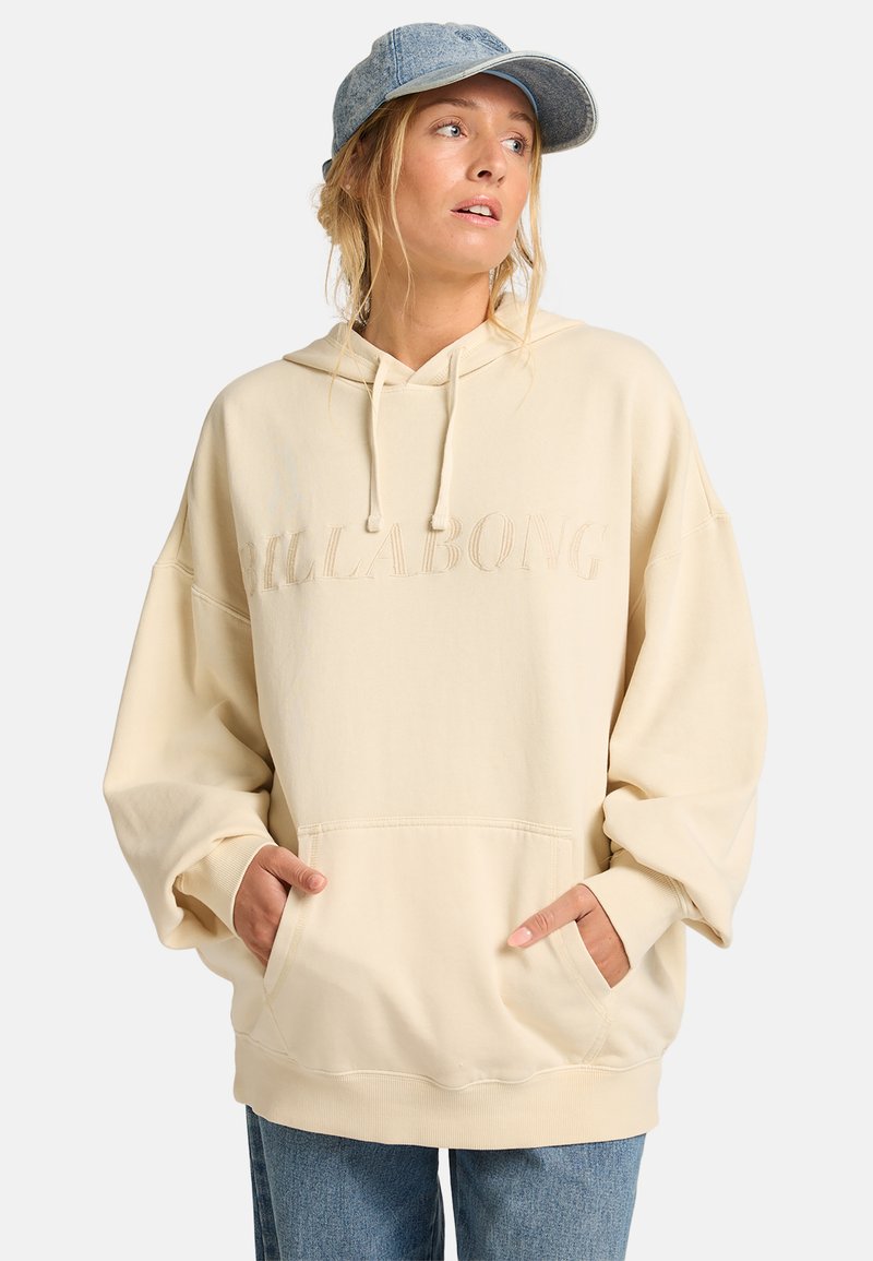 Creme-kleurige oversized hoodie met een textuur logo, een capuchon met trekkoord en een kangoeroezak. Gecombineerd met lichtblauwe jeans en een denim pet.
