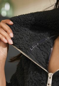 Veste zippée en polaire noire et duveteuse avec une surface texturée et une fermeture éclair argentée, montrant la doublure intérieure avec une étiquette contrastante.