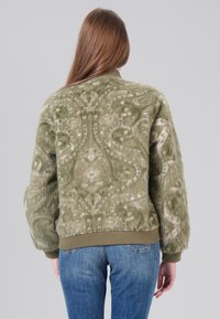 Veste bomber en fausse fourrure verte avec un motif en spirale texturé, des poignets et un ourlet côtelés, associée à un jean en denim bleu.