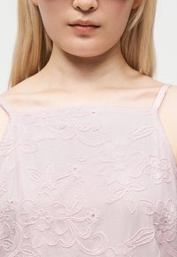 Femme à la peau claire et aux longs cheveux blonds portant un haut sans manches rose pâle brodé de fleurs, vue du menton à la clavicule.