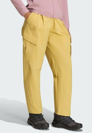 Persona que lleva pantalones cargo amarillos con grandes bolsillos laterales, zapatos de senderismo negros y una chaqueta rosa claro con cremallera, de pie con las manos en los bolsillos.