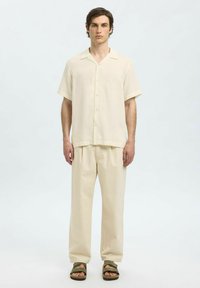 Camicia in lino a maniche corte di un beige chiaro, con bottoni, abbinata a pantaloni larghi dello stesso colore. Sandali con doppie cinghie in verde oliva.