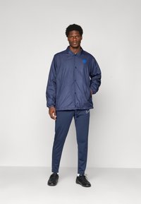 Man standing wearing a navy blue windbreaker jacket with logo, matching joggers, white socks, and black sneakers against a plain light background.
