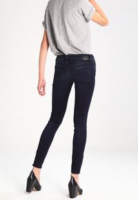 Skinny jeans in dark blue denim with subtle stitching. Paired with a gray T-shirt and black high-heeled shoes. Rear view displayed.