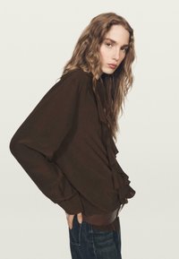 Brązowa bluzka z falbankami z przodu, długimi rękawami i luźnym krojem, w zestawieniu z ciemnymi dżinsami.