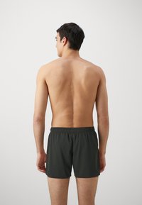 Shorts de bain vert foncé avec une taille élastique, fabriqués en matériau léger. Le modèle est vu de dos, sans chemise.