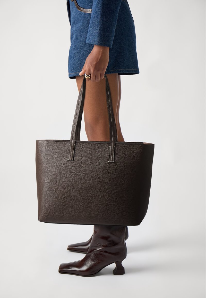 Personne portant une jupe bleue et des bottes brun foncé, tenant un grand sac cabas en cuir brun foncé par ses poignées.