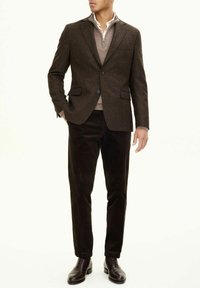 Blazer marrone in tweed con pattine, abbinato a un maglione beige chiaro con zip e pantaloni scuri in corduroy. Scarpe in pelle marrone.