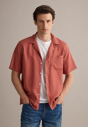 Joven con pelo corto y oscuro que lleva una camisa de botones color óxido de manga corta sobre una camiseta blanca y vaqueros azules, con las manos en los bolsillos.