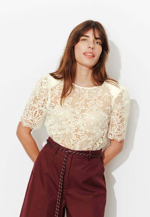 Femme aux longs cheveux bruns portant un haut en dentelle florale crème et un pantalon taille haute bordeaux avec une ceinture en corde, debout devant un fond blanc.