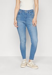 Pantalones vaqueros ajustados de talle alto azul claro en tejido de mezclilla con cinco bolsillos, que cuentan con un cierre de botones y ligeros detalles de desgaste. Combinados con zapatillas beige.