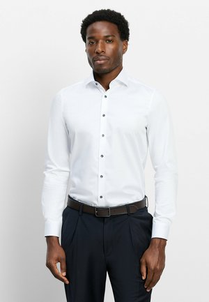 Mann trägt ein weißes langärmliges Hemd mit Knöpfen, schwarzen Gürtel und marineblaue Hose, steht vor einem schlichten hellen Hintergrund.
