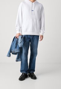 Sudadera blanca con capucha y un logo, jeans de mezclilla azules y zapatos negros gruesos; sosteniendo una chaqueta de mezclilla con la otra mano.