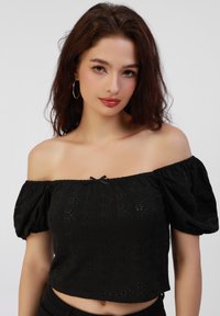 Sort cropped off-the-shoulder top med puffede ærmer, der har et struktureret stof med øjenlågsmønstre og en lille sløjfeaccent ved halsudskæringen.