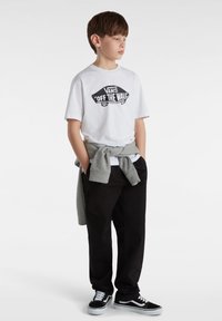 Witte katoenen t-shirt met zwart "VANS 'OFF THE WALL'" graphic, grijze sweatshirt om de taille geknoopt, zwarte broek en zwarte sneakers met witte accenten.