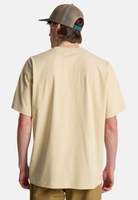 Μπεζ βαμβακερό μπλουζάκι με κοντά μανίκια και στρογγυλή λαιμόκοψη. Φορώντας ένα ανοιχτό καφέ δικτυωτό καπέλο με σκοτεινό γείσο και ρυθμιζόμενο πίσω λουράκι.