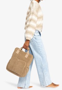 Borsa a tote beige con texture, dotata di due manici superiori, tasca frontale con zipper e dettaglio del logo, abbinata a jeans rilassati azzurri e un maglione a righe crema.