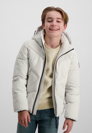 Witte gewatteerde jas met capuchon, voorzien van een geborduurd ontwerp, zwarte rits en contrasterende manchetten. Draagt over een lichte trui en blauwe jeans.