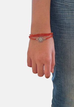 Mano con bracciale avvolgente di perline rosse e ciondolo geometrico in argento, appoggiata accanto a jeans di denim blu.