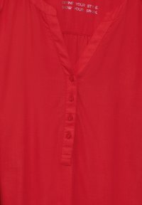 Chemise rouge à boutons avec un col en V, présentant une texture lisse et cinq boutons sur le devant. Inclut une étiquette imprimée à l'intérieur du col.