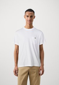 T-shirt en coton blanc avec un col rond et des manches courtes, arborant un petit logo vert sur la poche gauche de la poitrine.