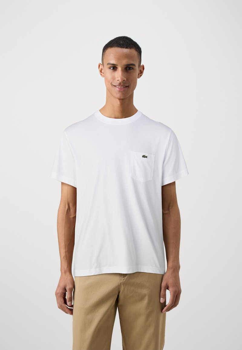 T-shirt en coton blanc avec un col rond et des manches courtes, arborant un petit logo vert sur la poche gauche de la poitrine.