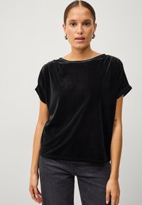 Sort fløjl kortærmet top med bred halsudsk�æring og opbrettede ærmer, der har en løs pasform og glat tekstur, parret med mørke jeans.