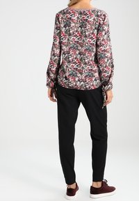 Mujer vestida con una blusa floral de manga larga, pantalones negros ajustados y zapatillas burdeos, de pie de espaldas sobre un fondo blanco.