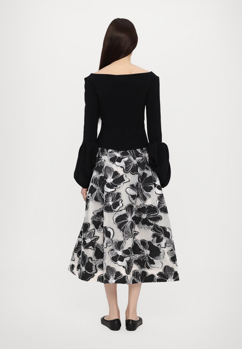 Top nero a spalle scoperte con maniche lunghe a campana, abbinato a una gonna di lunghezza media con un motivo floreale in nero e argento.