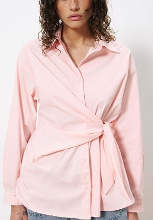 Chemise enveloppante rose clair avec des manches longues, fermeture à boutons sur le devant et un détail de nœud à la taille. Fabriquée en tissu doux et lisse avec une coupe ample.