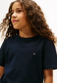 Ragazza con capelli ricci che indossa una maglietta nera con un piccolo logo rettangolare sul petto, che guarda leggermente di lato su uno sfondo semplice.