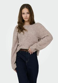 Pull rose pâle texturé à manches longues ; col arrondi. Porté avec un jean en denim foncé taille haute et une ceinture marron.