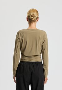 Duga majica s dugim rukavima u maslinasto zelenoj boji, izrađena od glatkog materijala, ima dizajn s naboranim leđima i kombinira se s crnim hlačama s elastičnim pojasom.