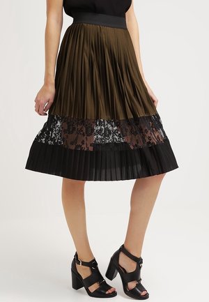 Vrouw die een geplooide midi-rok draagt met een olijfgroene bovenzijde, een zwart kanten middengedeelte, een zwarte zoom en zwarte sandalen met hoge hakken en enkelbandjes.
