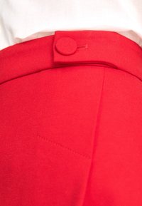 Jupe rouge présentant une texture lisse, une ceinture avec une fermeture par bouton rond, et un détail de poche cousue sur un côté.