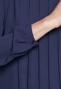 Blusa blu navy con dettagli a pieghe e polsini con bottoni. Tessuto liscio con un leggero effetto lucido, messo in evidenza da accenti sartoriali.
