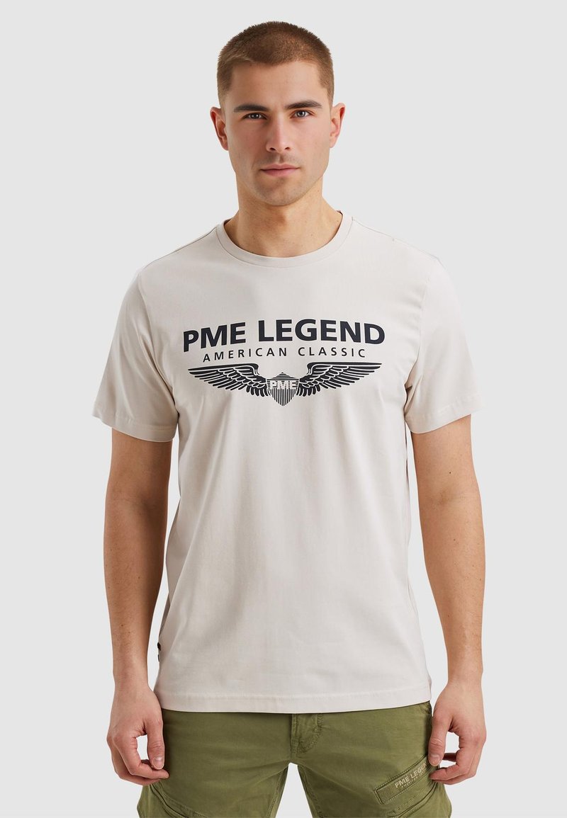Svetlo bež bombažna majica s črnim napisom "PME LEGEND AMERICAN CLASSIC" in grafiko krila, okroglo izrezana, kratke rokave in običajen kroj.