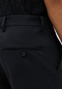 Κοντινό πλάνο μαύρου υφασμάτινου παντελονιού με κουμπωτή πίσω τσέπη και διακριτικό λογότυπο κοντά στη μέση στο ισχίο ενός ατόμου.