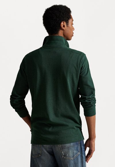 Groene gebreide pullover met een hoge kraag, lange mouwen en aansluitende manchetten. Gladde textuur en eenvoudig ontwerp. Gekocht met blauwe denim jeans.